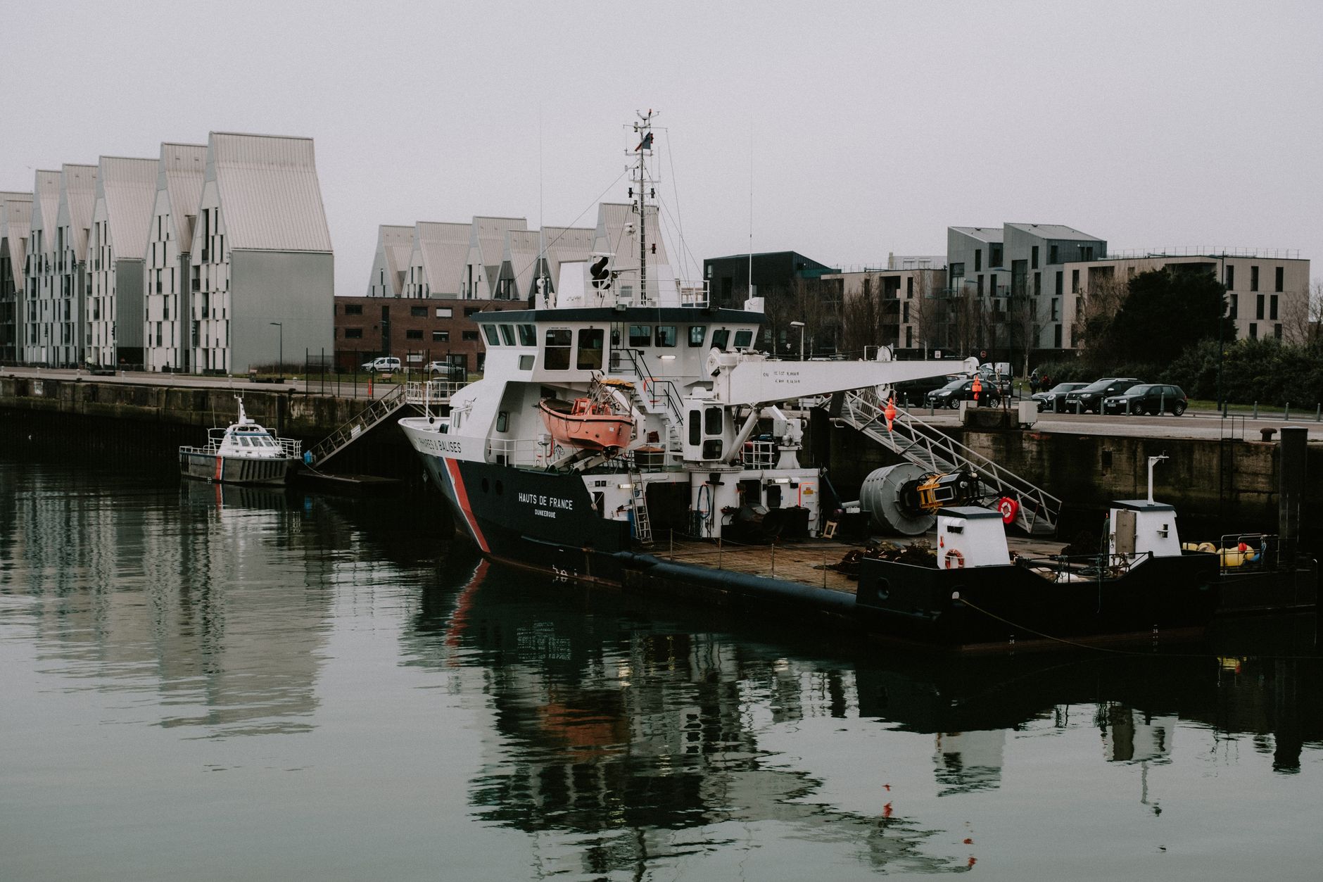 a sleek unmanned surface vessel moored at a coastal test facility, viewed from a low dock-level angle, with calm water reflecting the hull