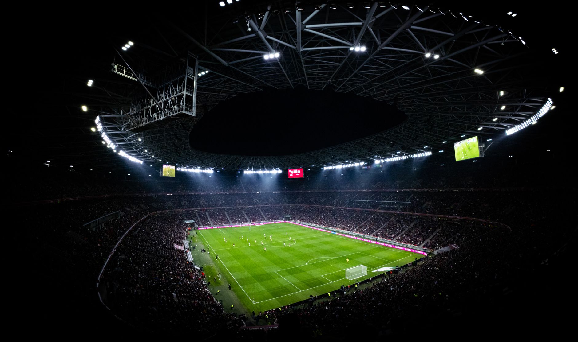 a largely empty professional football stadium interior at midday, showing the field turf markings, goal posts, and tiered seating with no crowd present