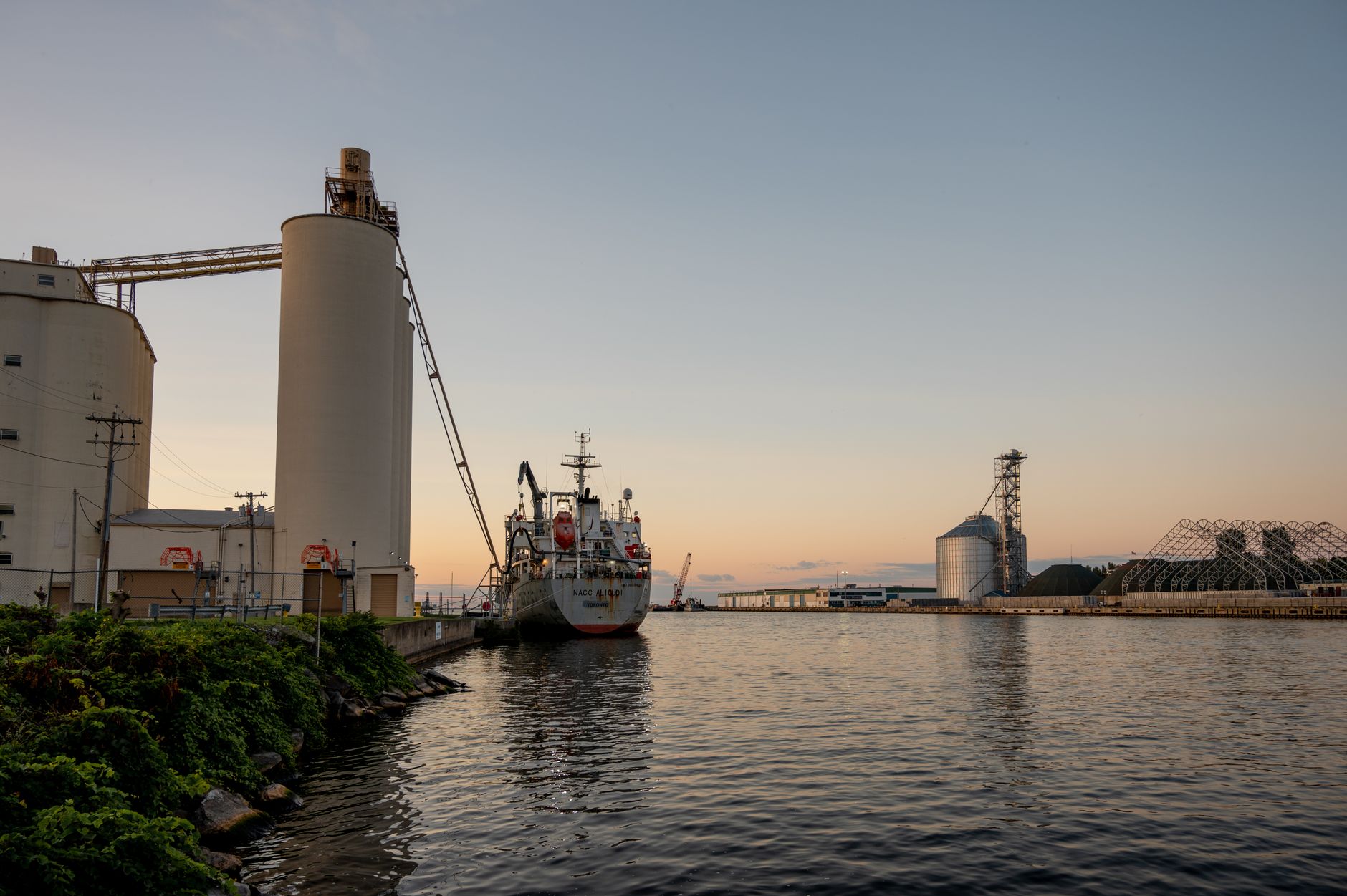 a commercial maritime port at dusk with a large cargo vessel at anchor, its hull and superstructure reflected in still water, photographed from a pier-level perspective
