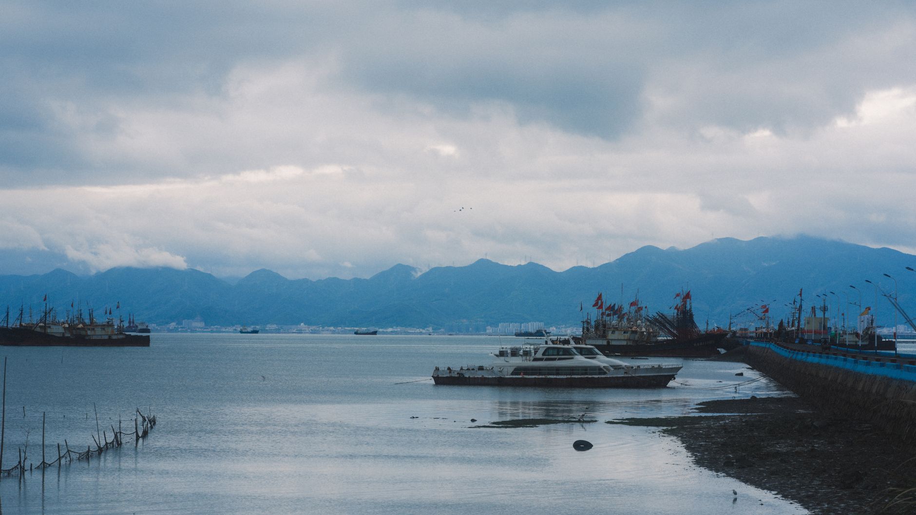 a sleek uncrewed surface vessel on calm open water during sea trials, viewed from a low angle at the bow, with a gray overcast sky and distant coastline