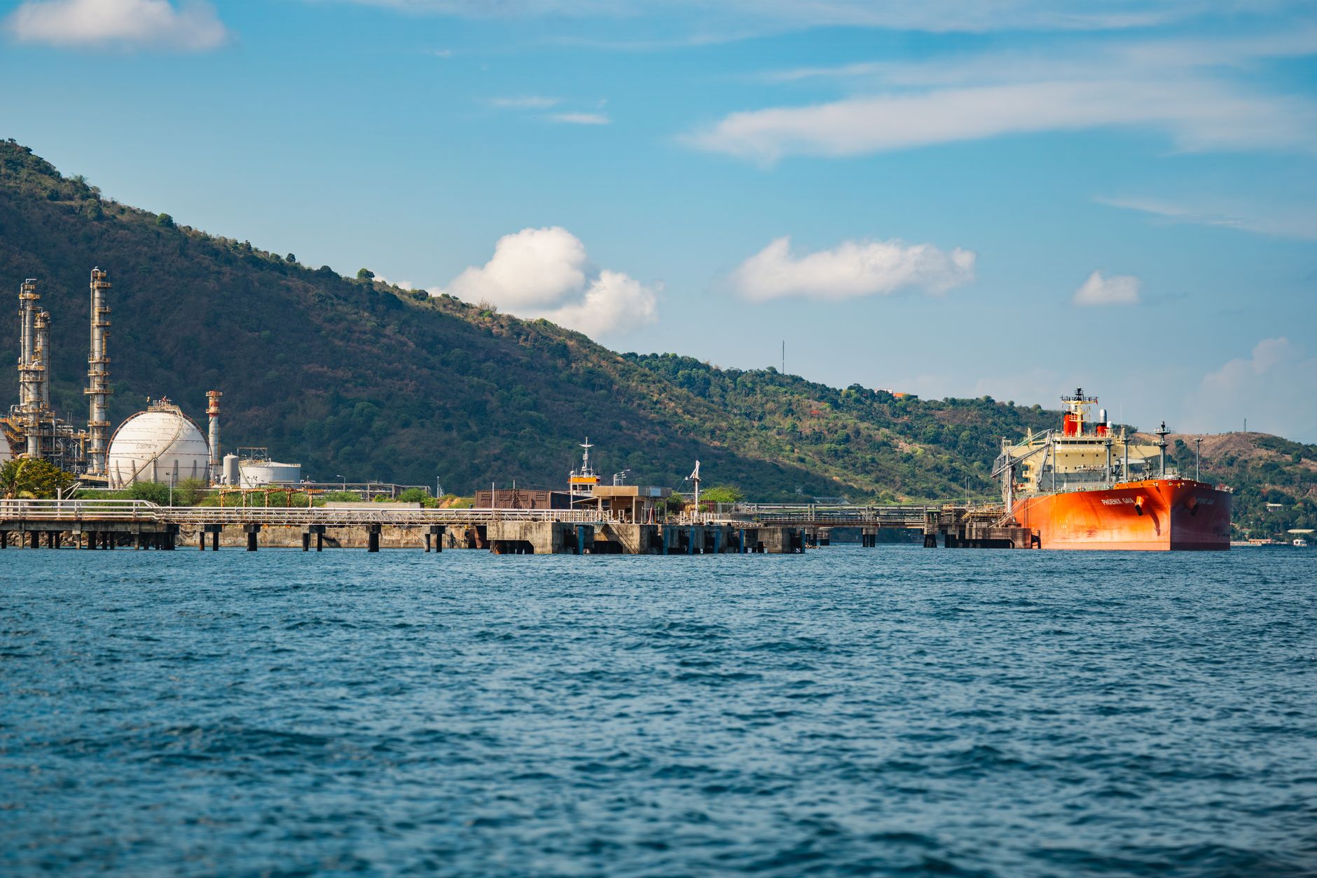 a large oil tanker transiting a narrow maritime chokepoint, viewed from an elevated coastal vantage point with arid hills in the background