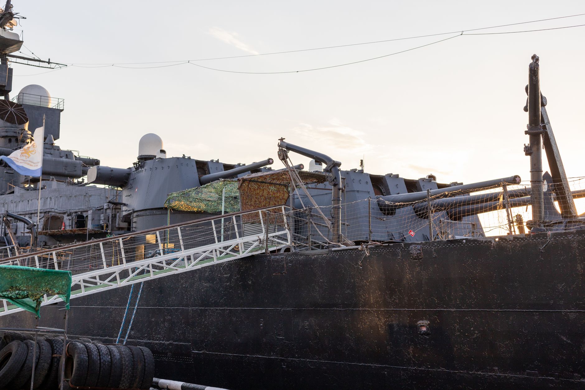 A compact naval vessel with a low-profile hull moored at a modern Nordic shipyard, surrounded by calm gray water and industrial dock infrastructure