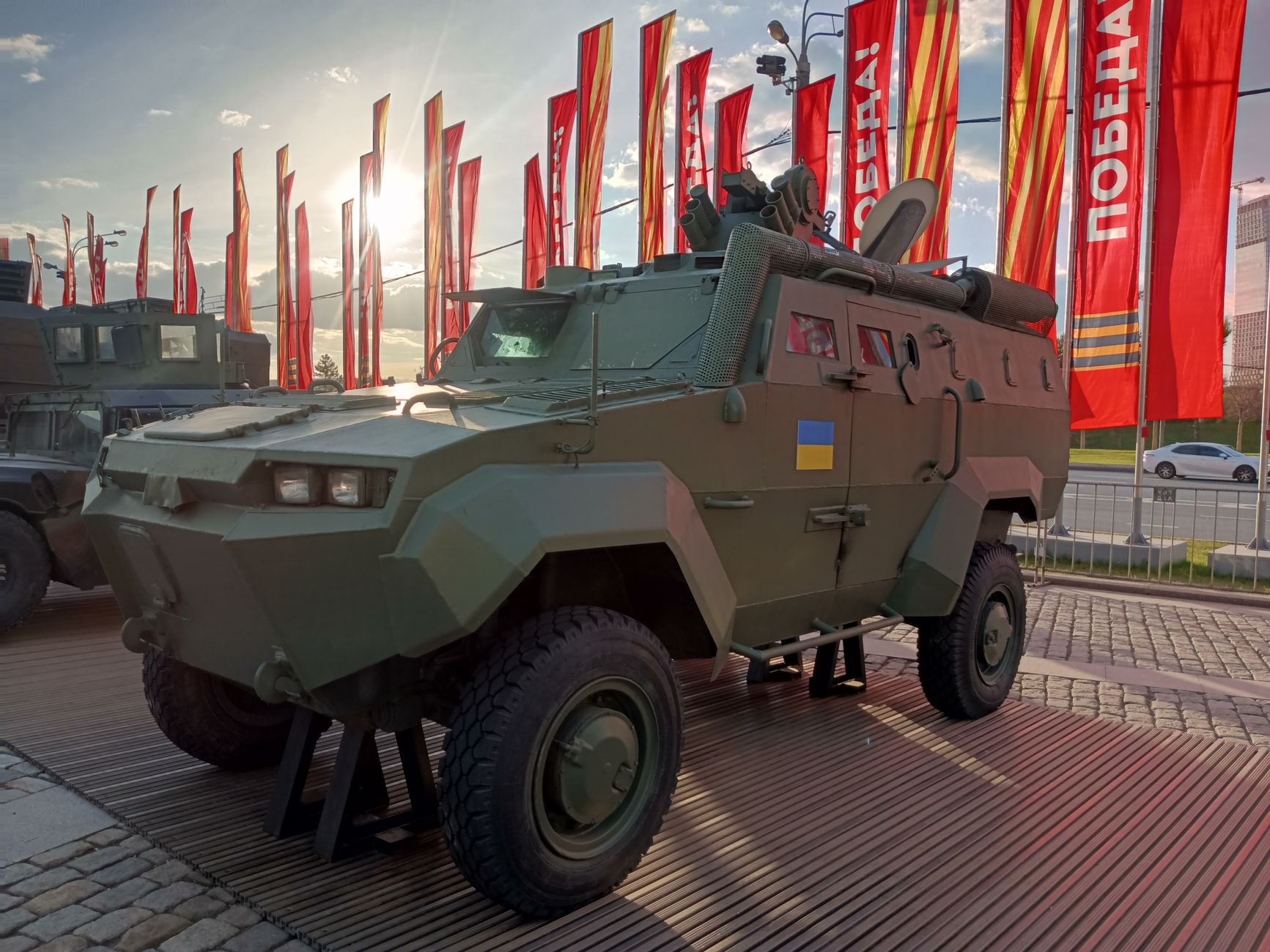 A row of armored military vehicles inside an open-sided field maintenance depot, viewed from ground level with workshop equipment and support vehicles visible in the background