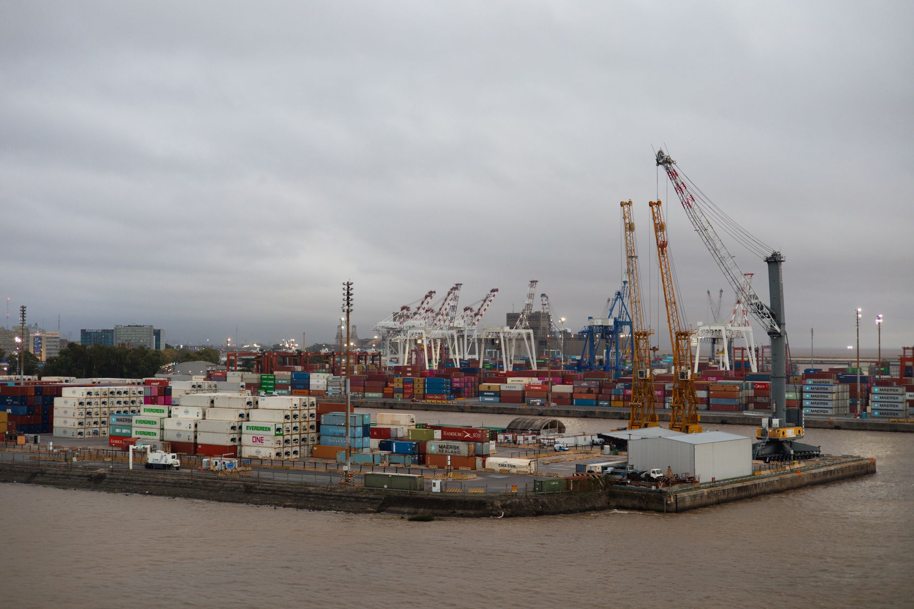 a large naval port facility viewed from above, with concrete quays, docked warships, and industrial cranes lining the waterfront under overcast skies