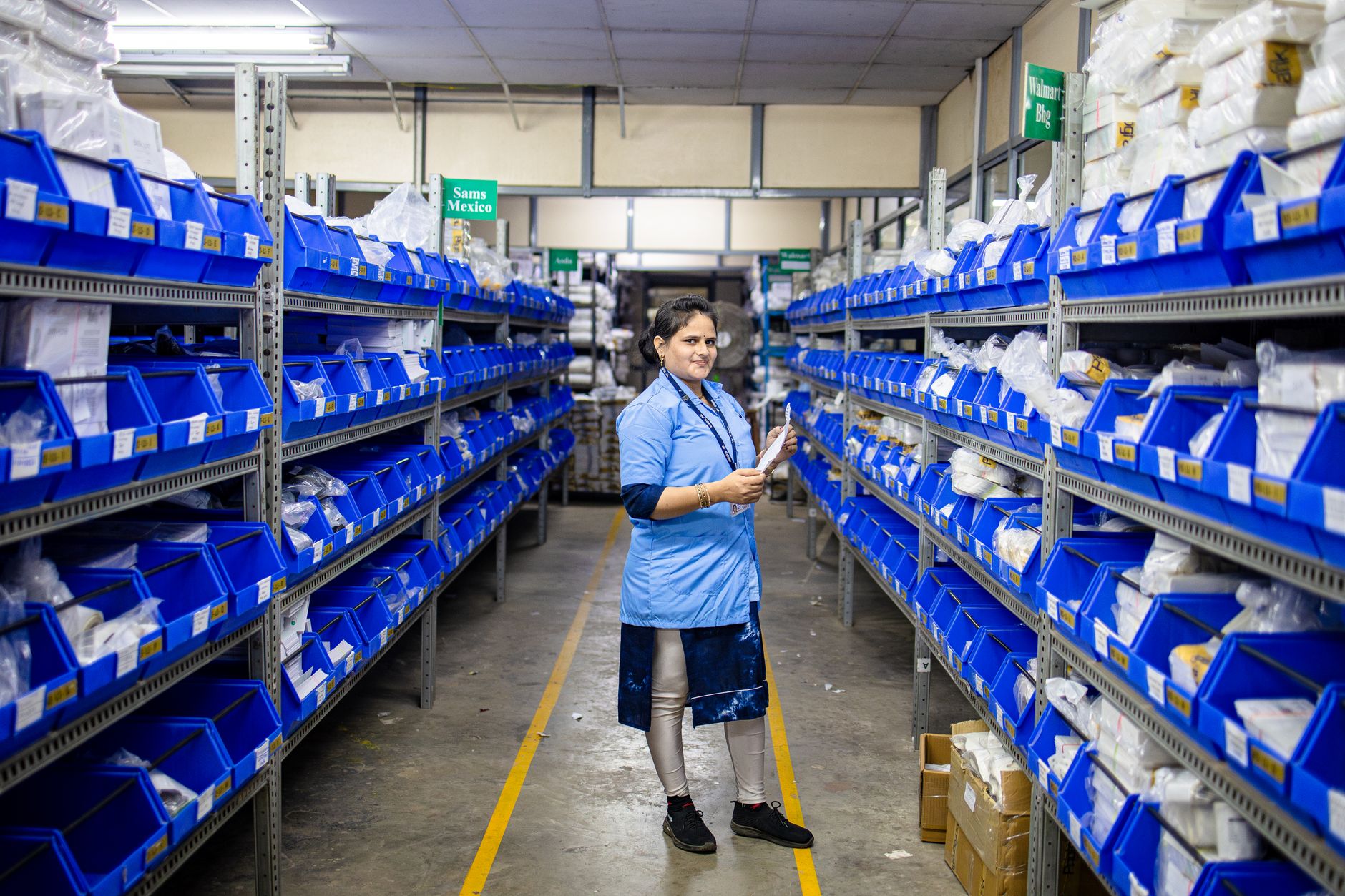 rows of cylindrical sonobuoy canisters arranged on industrial shelving inside a manufacturing facility, with overhead lighting and quality-control signage visible in the background