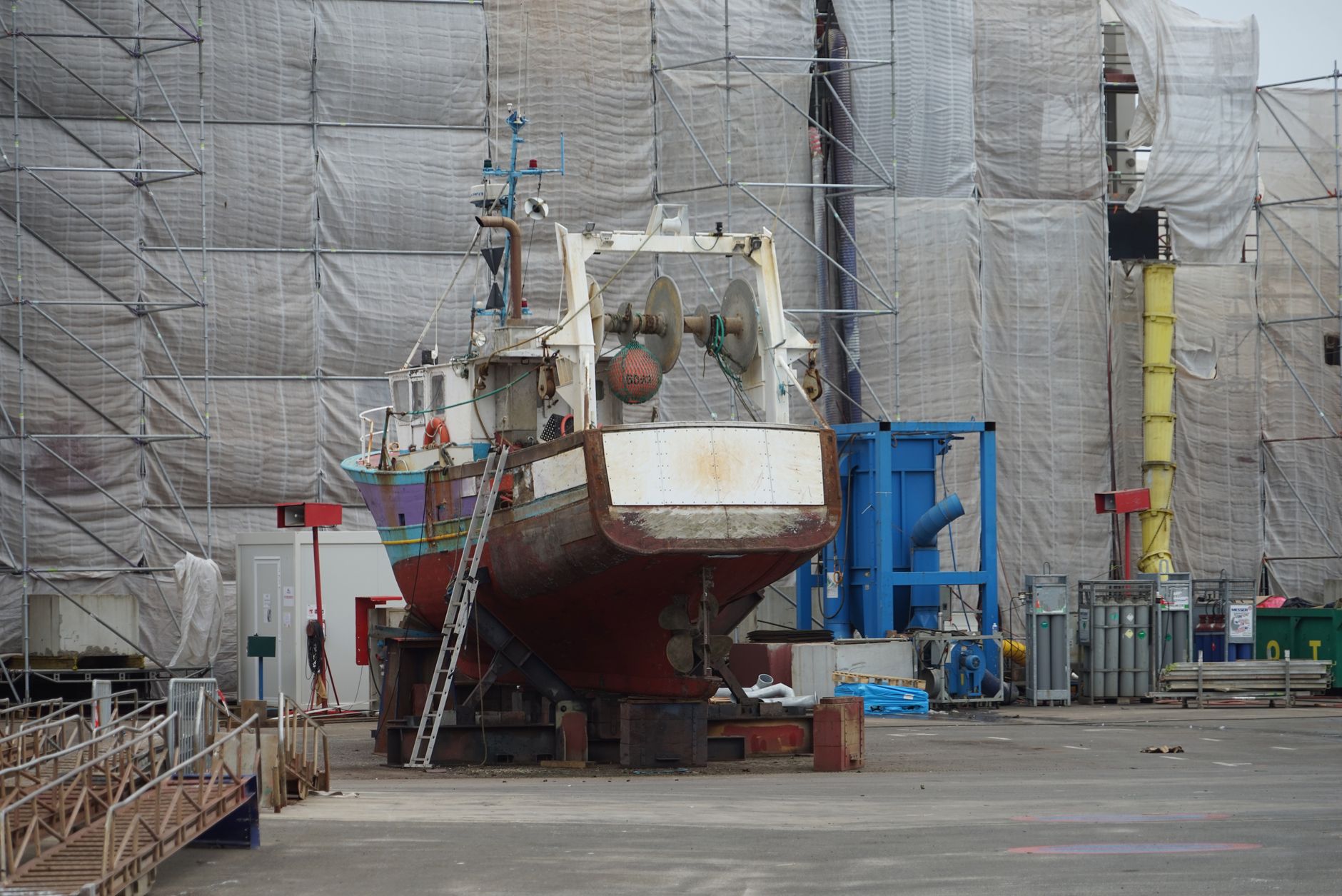 a naval shipyard dry dock with a frigate-sized hull under construction, scaffolding and industrial equipment visible throughout the facility