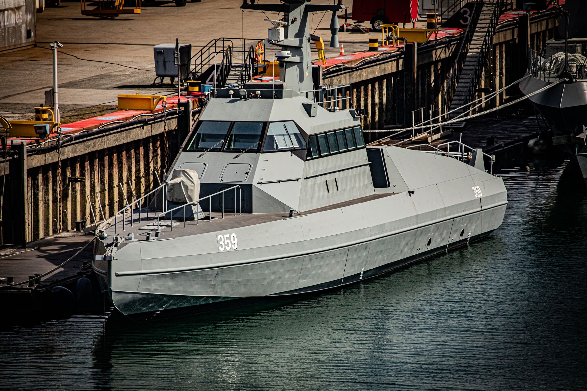 a naval research vessel at a pier with sonar equipment arrays visible on the deck, photographed from a dockside angle in overcast light