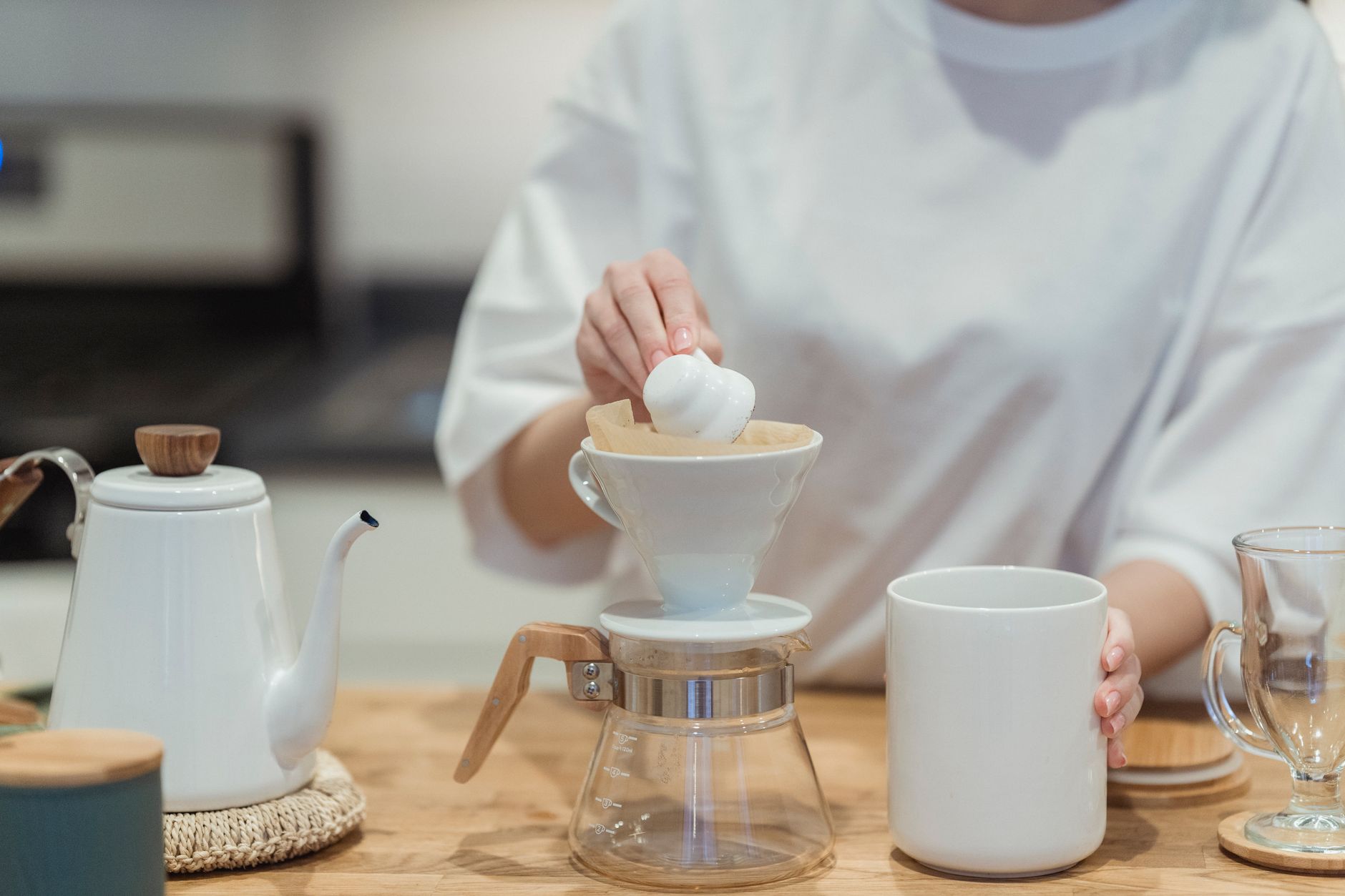 a close-up of a glass pour-over coffee dripper on a wooden countertop, with a ceramic cup beneath catching dark liquid, morning light through a kitchen window in the background