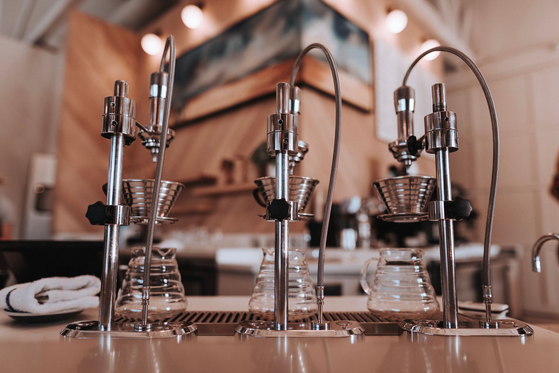 a row of standard drip coffee machines and paper filter cones arranged on a stainless steel counter in a large institutional kitchen, overhead fluorescent lighting, no people visible