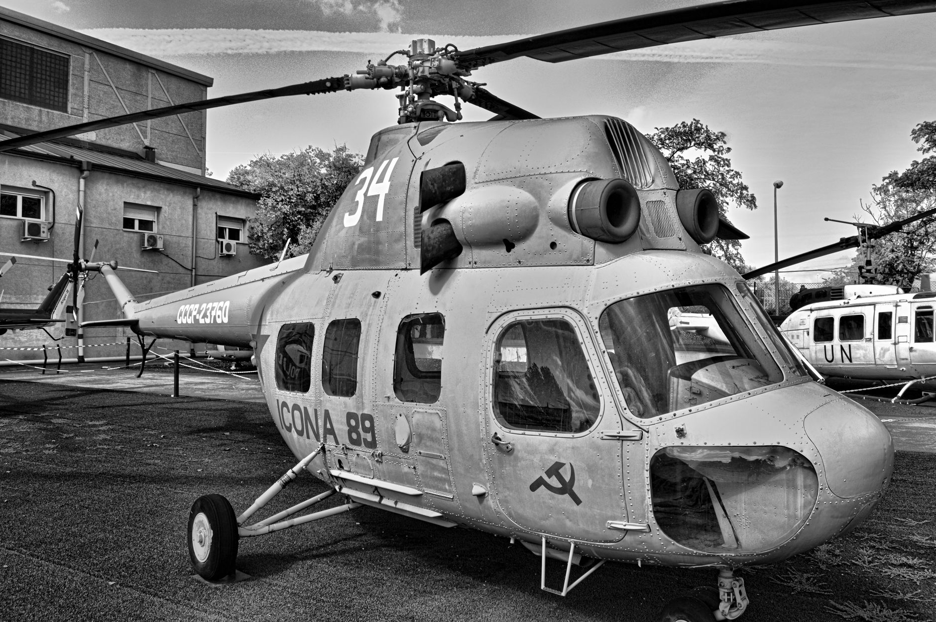 a massive twin-rotor Mi-26 heavy-lift helicopter on a sun-lit air base tarmac at an outdoor defense exhibition, viewed from the side to show its full fuselage length
