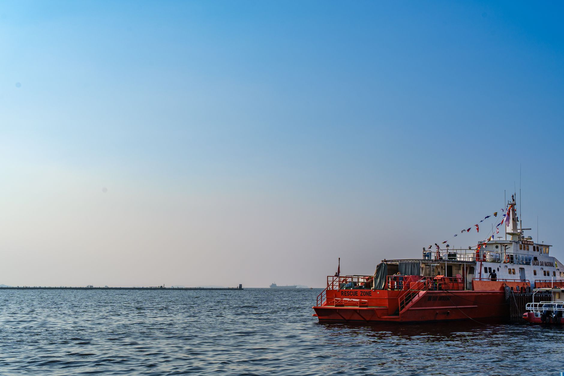 an unmanned surface vessel underway on open water, viewed from a slight elevation, with a naval base visible distantly on the horizon