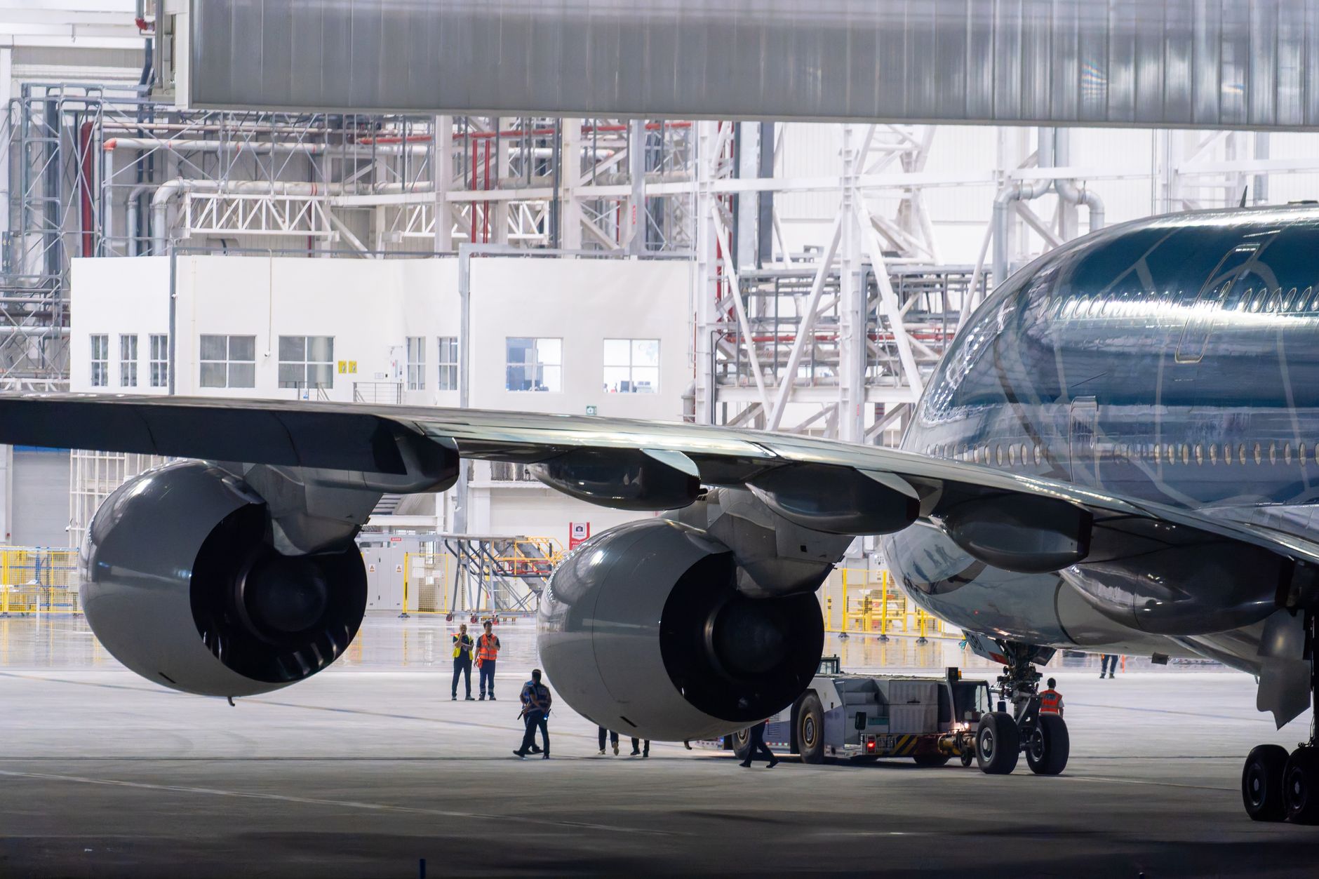 a ground-level view inside a large military aircraft maintenance hangar, with the massive wing and engine nacelles of a turboprop bomber visible on jacks, technicians and equipment visible in the background as small incidental figures