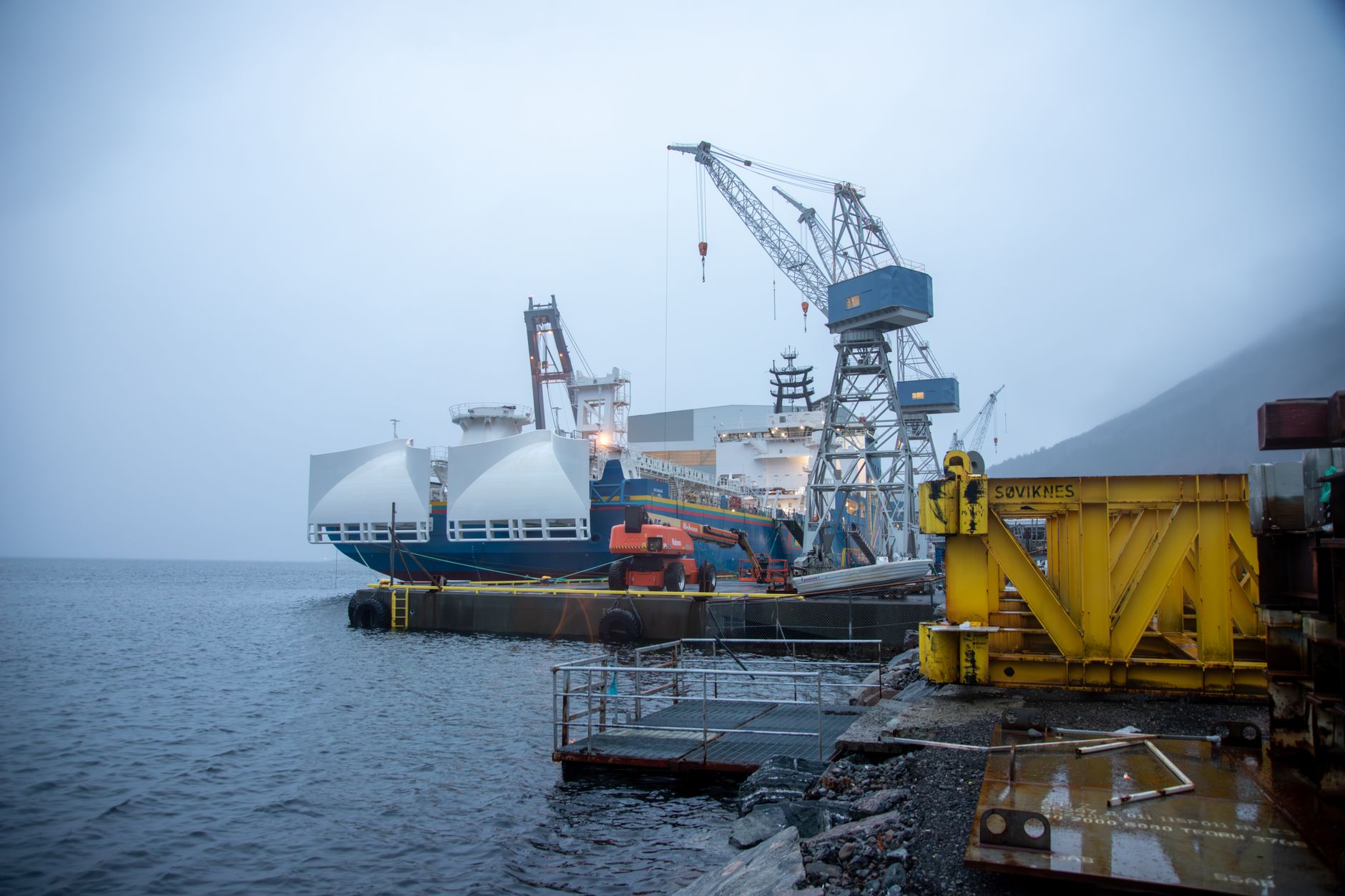 a newly launched offshore patrol vessel resting in calm harbor water alongside a large modern shipyard facility, surrounded by cranes and industrial infrastructure